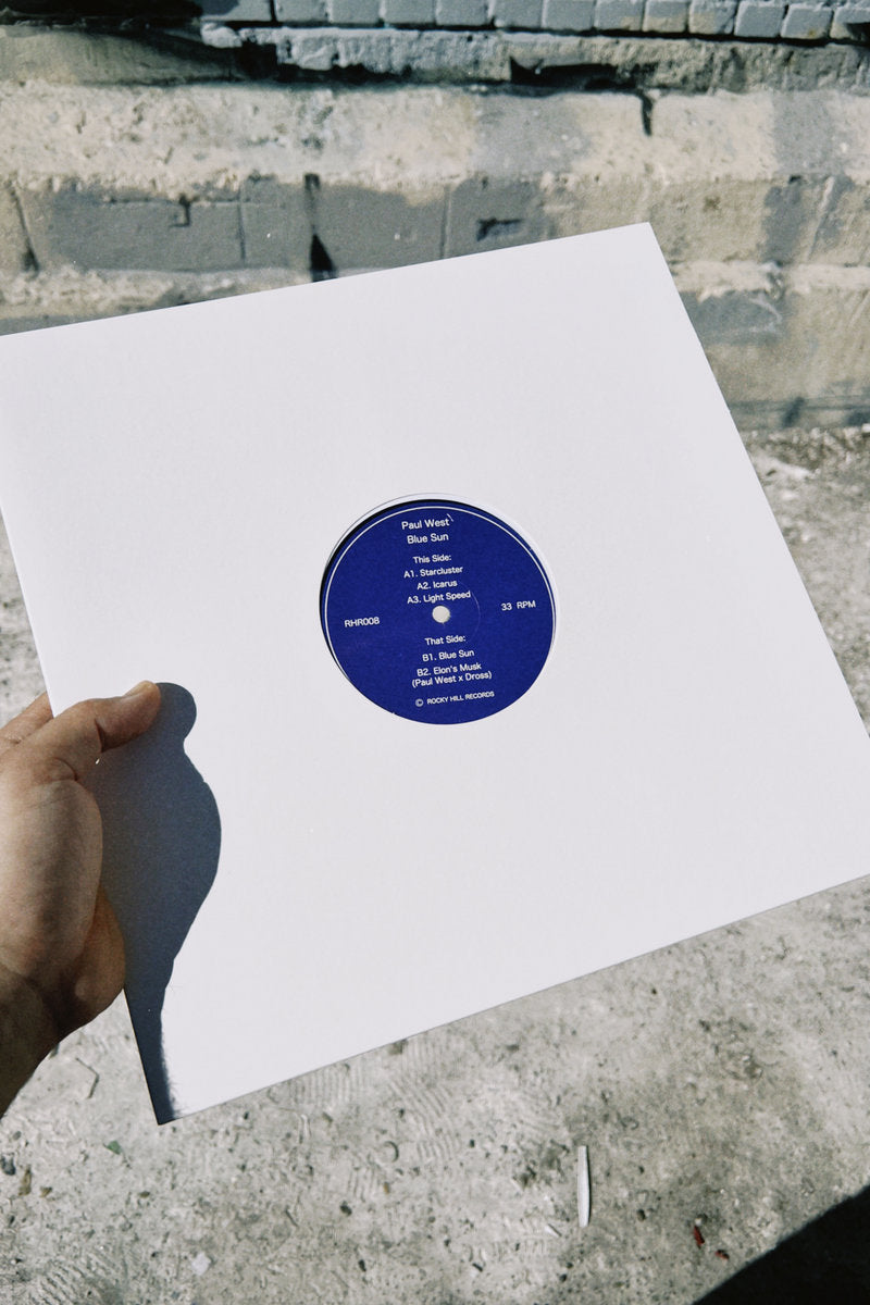 Portada del vinilo Paul West – Blue Sun 12" EP Rocky Hill, funda blanca minimal con label azul centrado sostenida en exterior urbano.