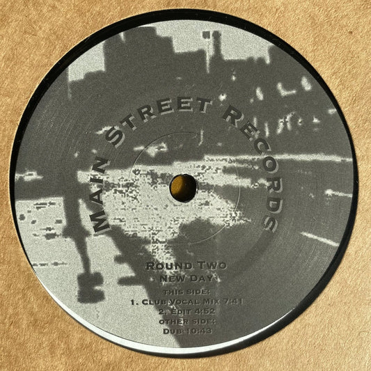 **Texto alternativo para la imagen "round-two-new-day.jpg":**
Etiqueta central del vinilo Round Two – New Day en su reedición 12" por Main Street Records, con diseño urbano en escala de grises y detalles de las pistas como "Club Vocal Mix" (7:41), "Edit" (4:52) y "Dub" (10:43).